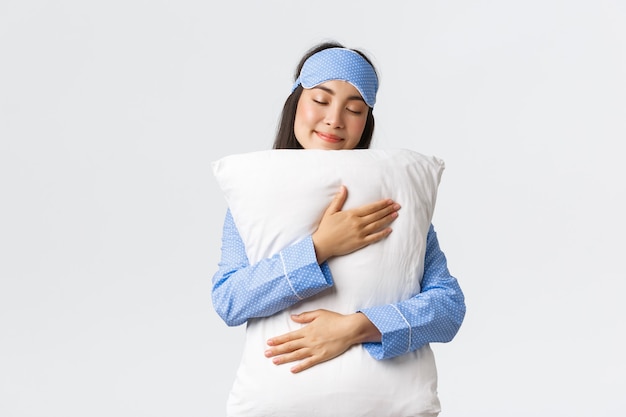 Free photo of a beautiful Asian girl smiling in pajamas and a sleeping mask, dreaming a sweet dream, hugging a pillow with a silly smile and closed eyes, standing on a white background, not wanting to wake up.